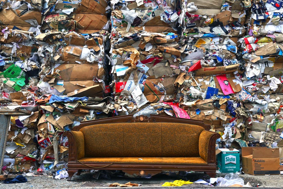 A vintage wooden sofa with ornate carved details and brown upholstered cushions is positioned in front of a large pile of mixed bulky waste, including flattened cardboard boxes, crumpled paper, and plastic packaging. The waste is stacked against a wall, filling most of the background and extending to the sides. The sofa is situated on a paved surface, likely outside a property or waste disposal area, with some scattered yellow and grey items nearby. The scene suggests the process of clearing out household items during a home relocation or furniture disposal, with the waste materials prepared for collection or disposal services. The overall image captures the clutter associated with large item removal, supported by [COMPANY_NAME]'s professional removals and waste disposal solutions, emphasizing the importance of proper planning for furniture transport and efficient bulk waste management.