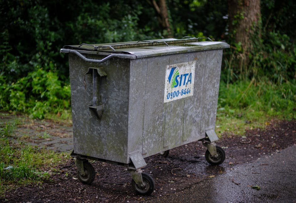 A large, grey, wheeled waste bin with a metal lid and a blue and green logo on the side, placed on a paved outdoor area beside a grassy verge with trees and shrubs in the background. The bin appears stained and weathered, typical of containers used for bulky waste disposal in residential areas. It is positioned near the edge of the pavement, adjacent to a dirt area and greenery, indicating it is waiting for collection or disposal as part of a home relocation or moving process. The wheels of the bin are visible, and the environment is lit with natural, overcast daylight, typical for outdoor loading or waste collection scenarios. The presence of this bin suggests discussions of practical waste management solutions associated with packing and moving services provided by companies like Man with Van Highams Park, supporting efficient relocation logistics.