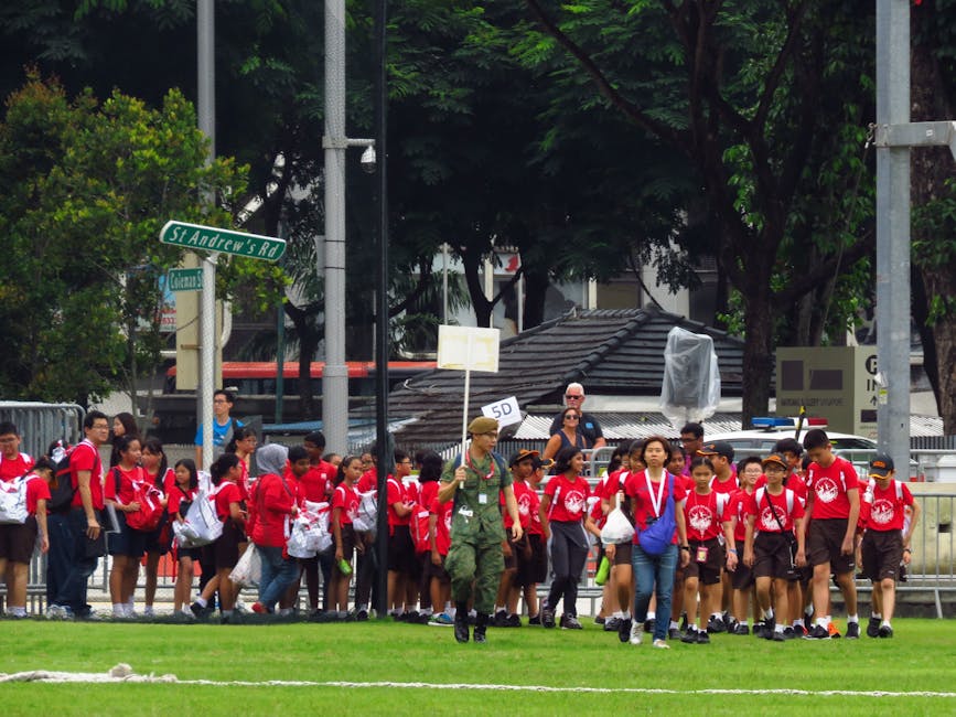 A group of school children dressed in red uniforms, carrying plastic bags and backpacks, are walking in a line on a grassy area near a street intersection marked by a street sign for St. Andrew's Road, under a canopy of large green trees. An adult in military-style clothing is walking alongside them, holding a sign with the number '5D'. In the background, there is a small building with a tiled roof, several street lamps, and parked cars, indicating an urban environment. The scene appears to be part of a school outing or organized activity, with the children preparing for a home relocation or part of a supervised moving or transport process, as managed by Man with Van Highams Park, specializing in removals and moving services.