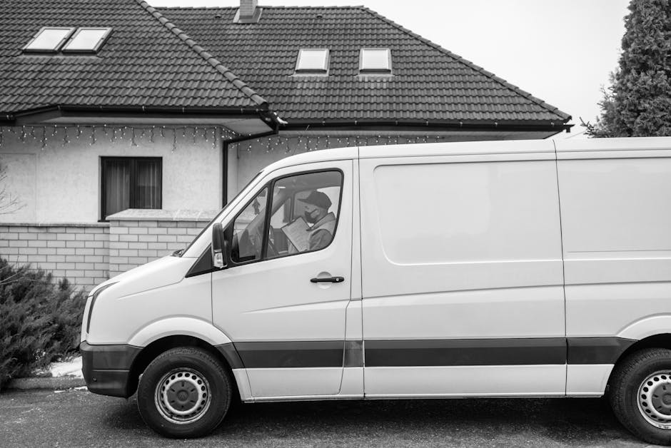A man wearing casual clothing, including a cap, is walking alongside a white panel van parked on a residential street. The van is positioned parallel to the pavement, with the sliding side door closed