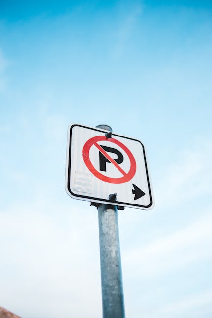 A street sign mounted on a metal pole against a clear blue sky indicating no parking with a red circle and diagonal line over a black letter 'P', and an arrow pointing right. The sign is located in an outdoor urban environment, situated near a property entrance, likely used to inform drivers about parking restrictions during house removals or moving activities in Highams Park. The sign's metallic post shows signs of weathering, and the background is free of other visual obstructions, emphasizing its visibility for passing motorists or pedestrians. This image reflects the importance of understanding local parking regulations when planning a home relocation with services such as those offered by Man with Van Highams Park, especially when undertaking furniture transport or loading and unloading processes related to moves, ensuring compliance with council rules and smooth logistics.