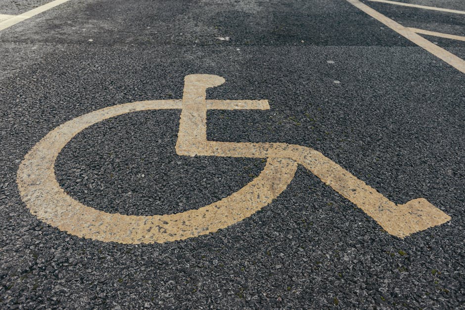 A close-up view of a paved parking space featuring a faded yellow wheelchair-accessible parking symbol painted on dark asphalt. The symbol consists of a circular shape representing the wheelchair's wheel, with a simplified figure seated in it, and a horizontal line indicating the footrest. The painting shows signs of wear, with some areas chipped and uneven, highlighting frequent use and weather exposure. In the background, the parking area has white lines marking individual spaces, with a portion of a vehicle visible at the upper edge of the image. This scene is typical of urban or residential parking facilities, relevant to house removals and moving services, as it illustrates an accessible parking space that might be used during a home relocation, carried out by professionals like Man with Van Highams Park, who handle furniture transport, packing, and loading processes.