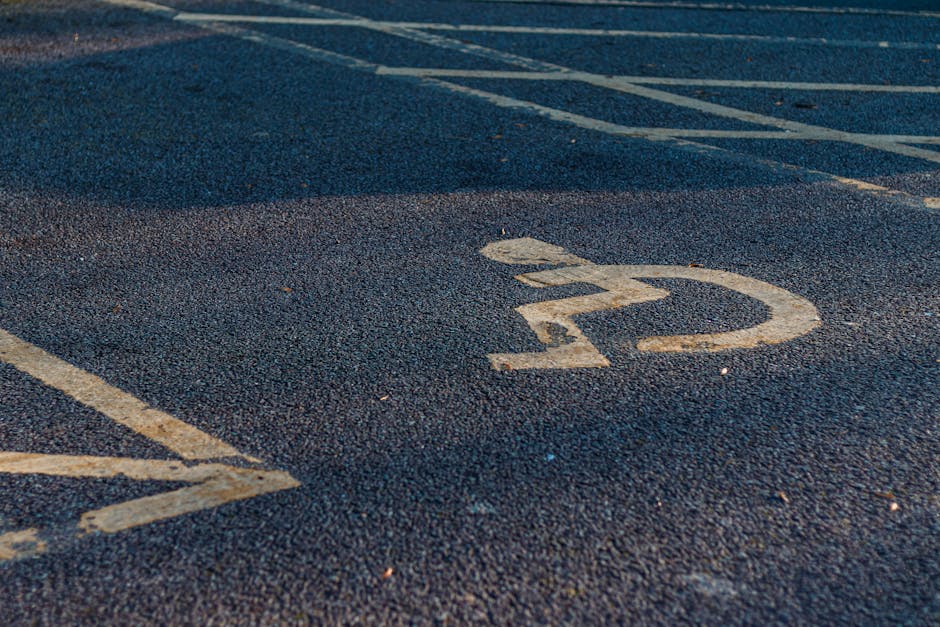 A close-up view of an asphalt parking lot surface featuring designated white-painted markings for accessible parking spaces, including the international wheelchair symbol within a rectangular outline. The markings are clear and well-maintained, with the surrounding area showing additional parking lines and space numbers. The textured asphalt provides a contrasting background to the clean, crisp white paint. This image illustrates the parking environment outside a residential or commercial property in Highams Park, pertinent to the planning of house removals or furniture transport, where parking permits and access considerations are essential. Man with Van Highams Park specializes in home relocation and moving services, ensuring suitable parking arrangements for a smooth loading and unloading process during house removals.