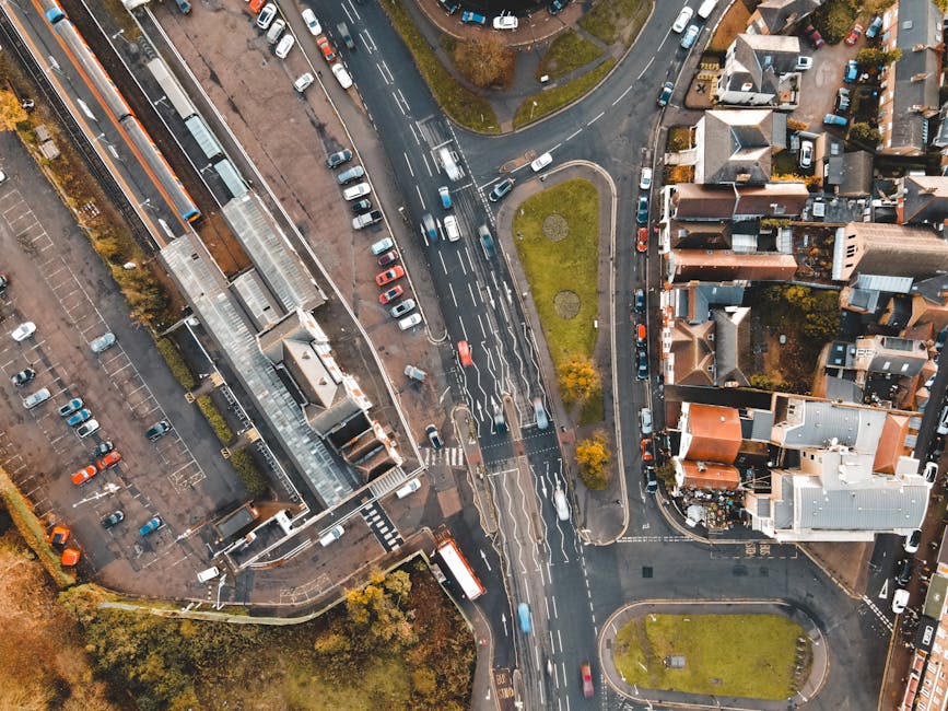 An aerial view of a busy intersection near Highams Park Station, showing multiple cars in motion on the road and parked along the curb, with a small green grass median strip in the center. Surrounding the junction are residential houses with varying roof styles and colors, a large multi-story building with a glass entrance canopy on the left side, and several parking lots partially filled with vehicles. The lighting appears to be bright and natural, indicating daytime. This scene illustrates the urban environment where Man with Van Highams Park might facilitate home relocation and furniture transport, highlighting the logistical context of moving and loading goods in a suburban setting.
