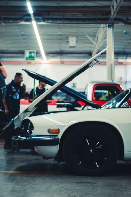 A close-up view of a white vintage car with its front hood and driver’s side door partially open, positioned inside a brightly lit indoor vehicle exhibition space. The car has black wheels and visible front headlights, with a glossy finish reflecting the overhead lighting. In the background, there are people observing the vehicle, some appearing to be engaged in conversation, alongside other vehicles and exhibition fixtures. The setting suggests a vehicle showcase or car event, which is relevant to the context of transport and loading processes involved in house removals or furniture transport, with the focus on the car as a key object in moving or relocation activities. The professional environment is well-lit, highlighting the details of the car and the space around it, aligning with the themes of vehicle loading and furniture transportation services provided by companies like Man with Van Highams Park.