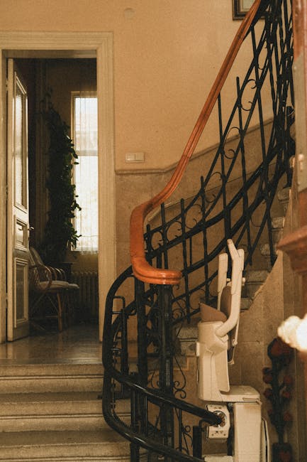 Inside a residential property, a staircase with a black wrought-iron railing and a wooden handrail curves upwards towards an upper floor. The staircase is located near a doorway, through which a room with a window, a couch, and some indoor plants is visible, illuminated by natural light. At the base of the staircase, a white home lift or stairlift is installed, with the folded seat resting on its platform. The area around the staircase appears tidy, with neutral wallpaper or painted walls, and a marble or tiled floor. The scene suggests a setting where furniture transport or home relocation services might involve navigating narrow or small spaces such as stairs or small lifts, a typical challenge addressed by [COMPANY_NAME] in conducting safe, efficient removals or packing and moving operations.