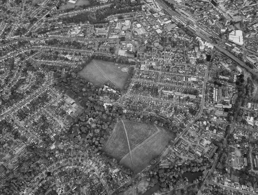 Aerial black-and-white photograph of a suburban area showing numerous residential houses, roads, and green spaces, including two large open fields with visible walking paths crossing through them. The fields are surrounded by trees and a mix of detached and semi-detached homes with back gardens. In the foreground, a row of houses lines a curved street, while the background reveals a more densely built area with terraced houses and commercial buildings. A railway line runs along the top edge of the image, with visible train tracks and station infrastructure. Natural light illuminates the scene evenly, and no people or vehicles are visible in the image. This landscape provides context for house removals or home relocation services, illustrating the environment where furniture transport and packing and moving activities may take place, as managed by Man with Van Highams Park.